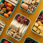 Top view of vibrant lunch boxes filled with assorted snacks on yellow background.